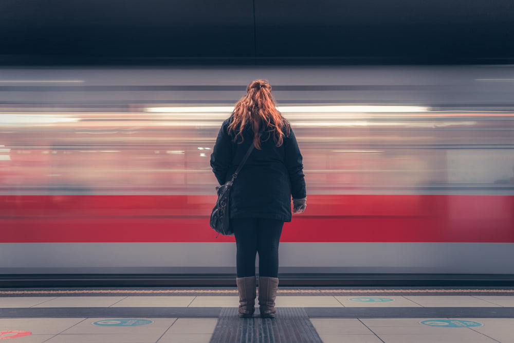 Young woman with wavy red hair stands alone as a subway train passes her in a blur